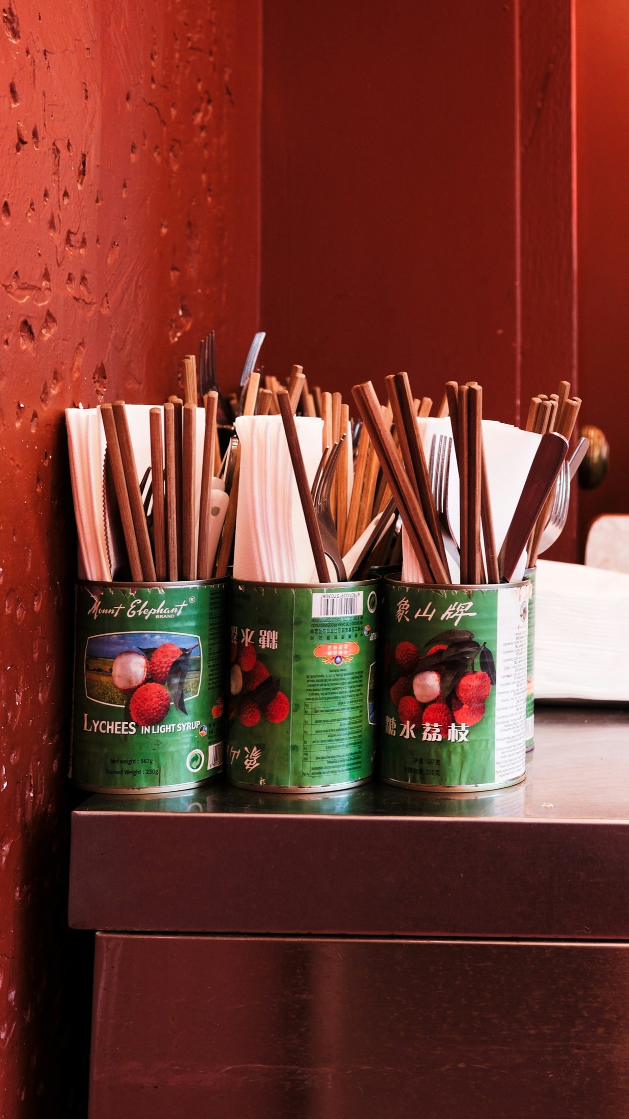 Chopsticks and cutlery held in reused lychee cans against a red wall in a Chinese restaurant